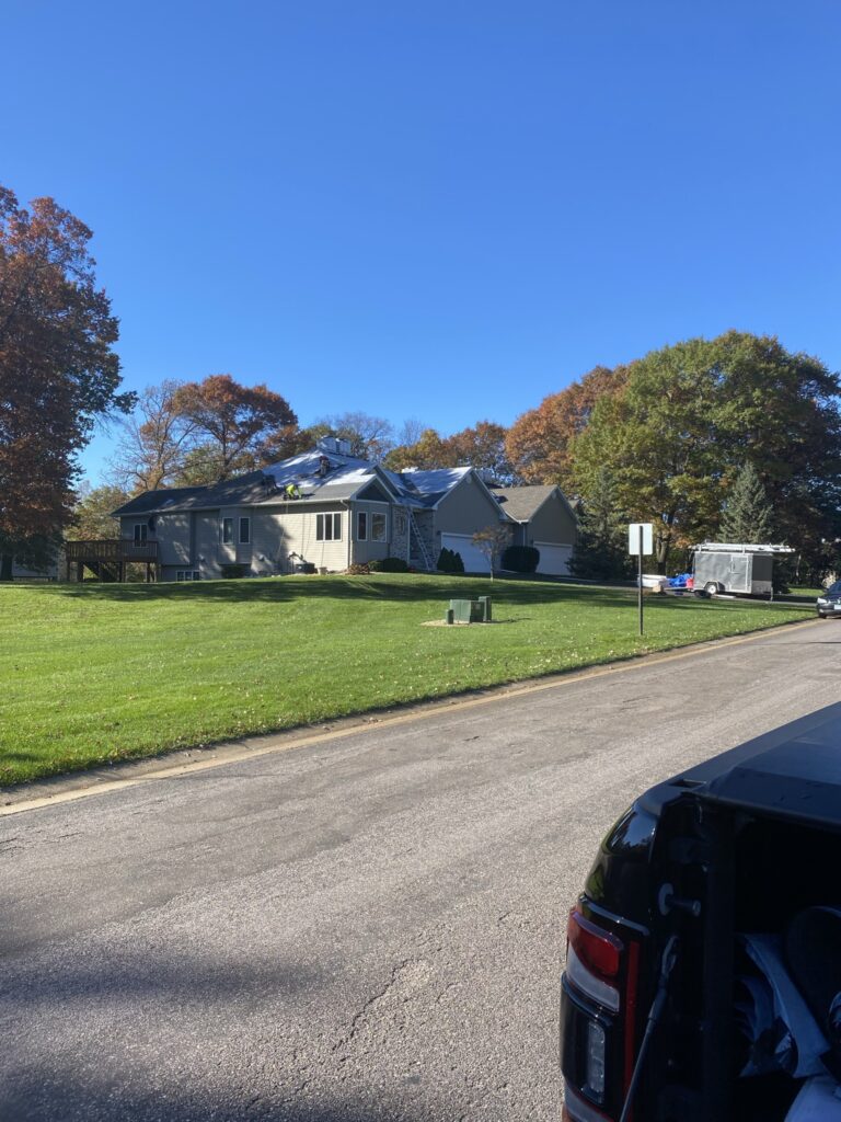 A suburban house with fall-colored trees in the background, sits on a large green lawn under a clear blue sky. Part of a black vehicle and a trailer are visible on the right side of the image.